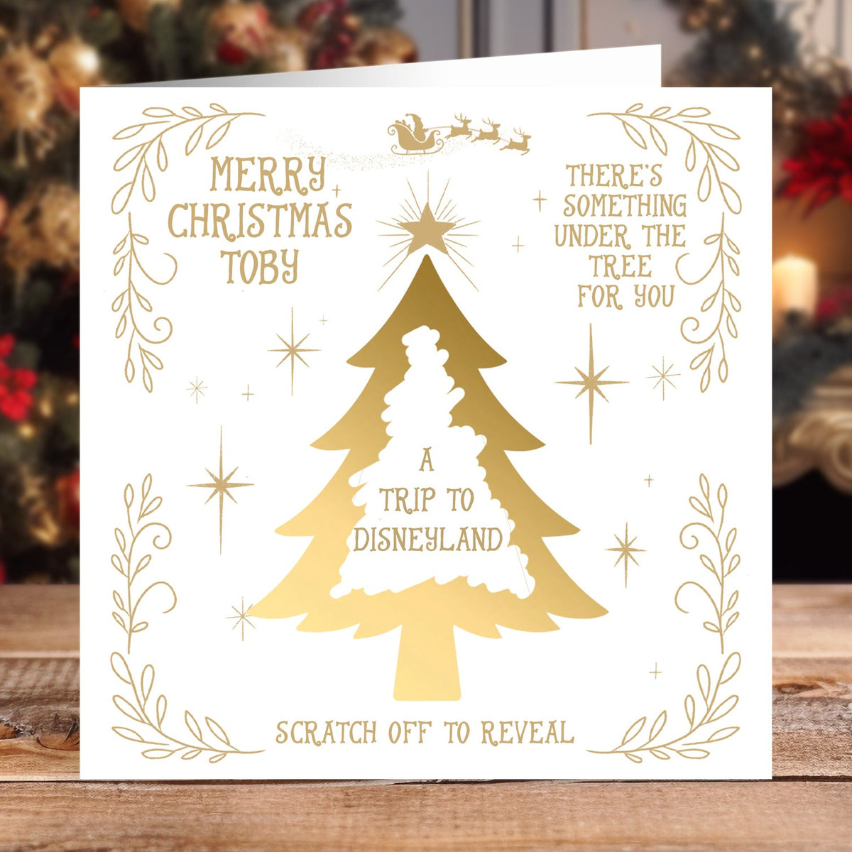A Christmas greeting card with gold foil lettering and graphics on a white cardstock, displayed on a wooden table with a blurred Christmas tree in the background. The card reads 'MERRY CHRISTMAS TOBY', 'A TRIP TO DISNEYLAND', and 'SCRATCH OFF TO REVEAL'.