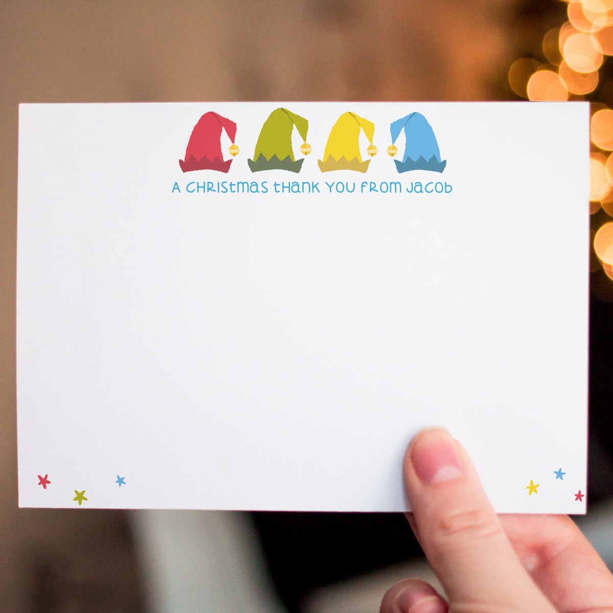 A hand holding a white personalized Christmas thank you card with colorful elf illustrations and the text 'A CHRISTMAS THANK YOU FROM JACOB' against a warm, blurred holiday background.
