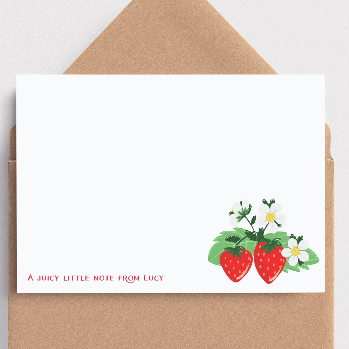 A white stationery note card with a small strawberry and flower illustration in the lower right and the text 'A JUICY LITTLE NOTE FROM LUCY' in red, shown with a brown kraft envelope on a light gray background.